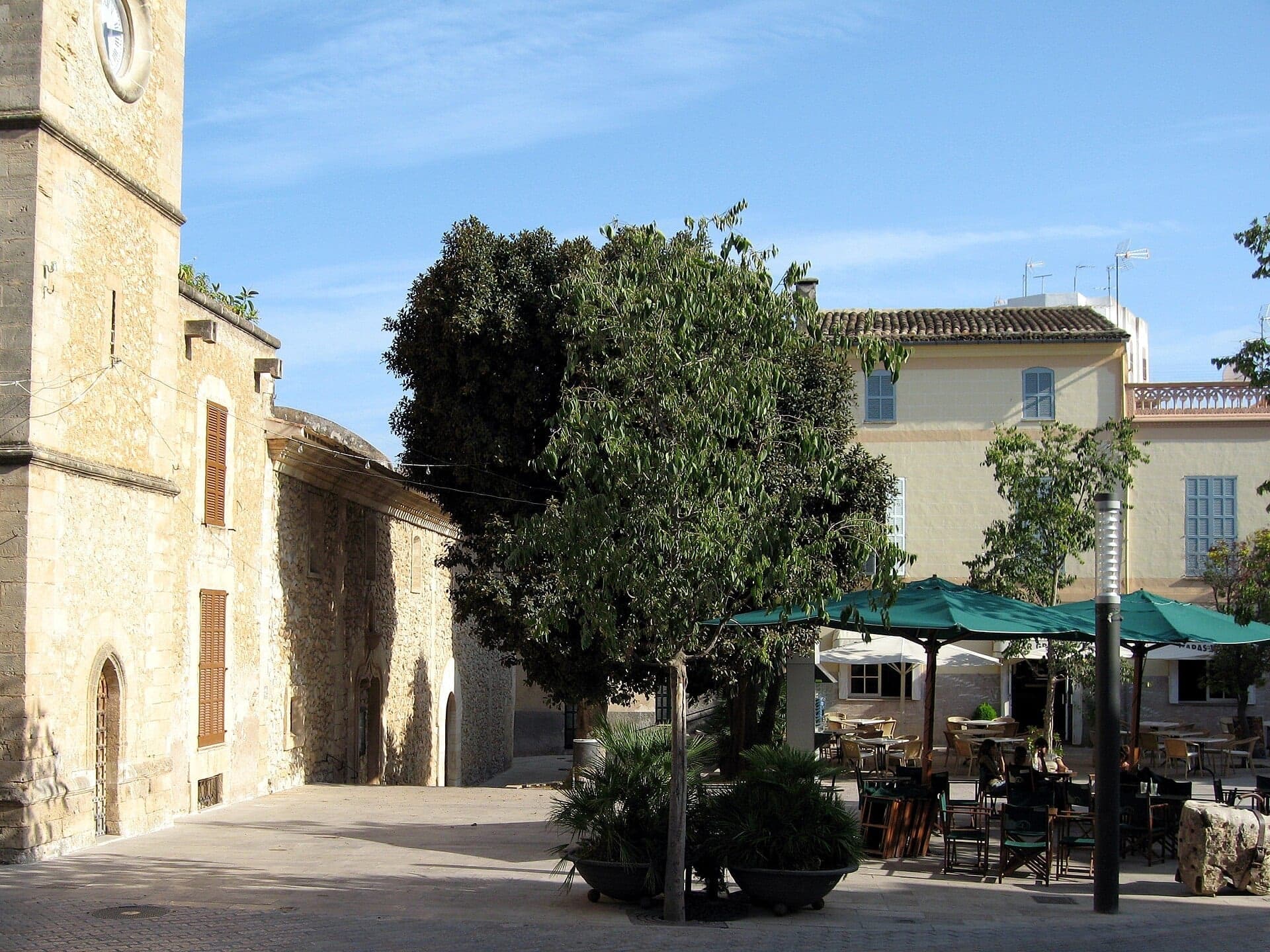 La plaza del casco antiguo de Son Servera, con la torre de piedra de la iglesia parroquial y una terraza de café a la sombra.
