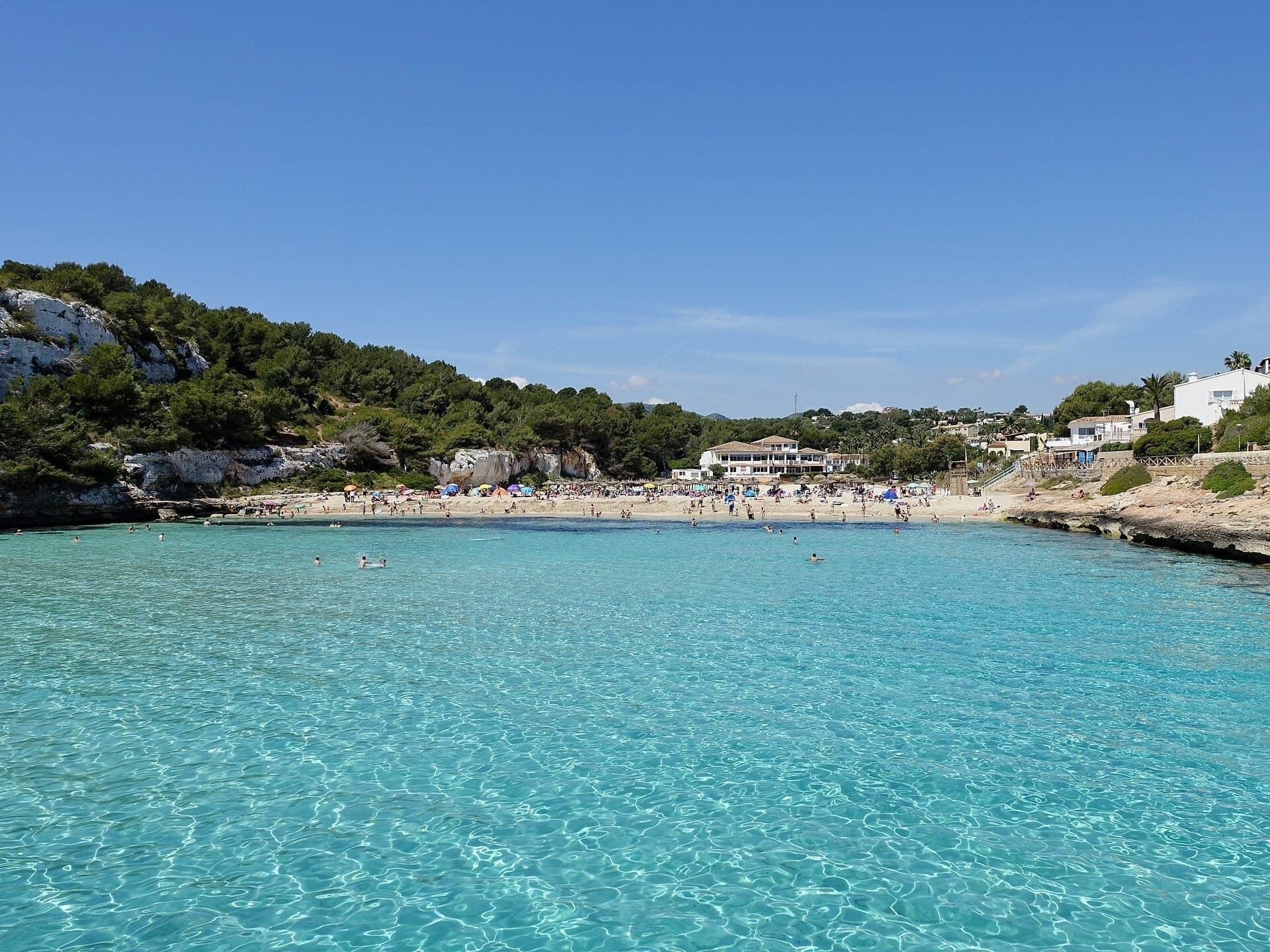 La cala turquesa y la playa de arena de Cala Romántica (S'Estany d'en Mas), entre acantilados con pinos.
